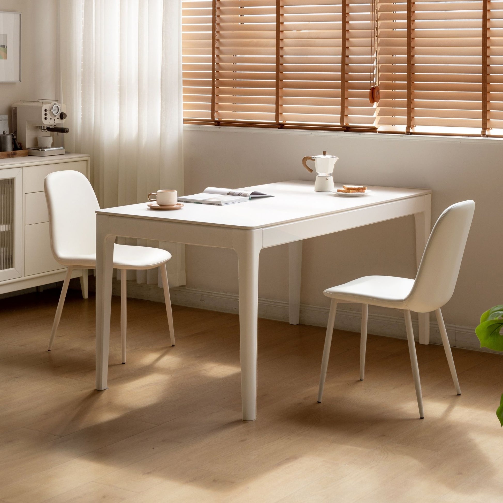White dining table with chairs in a room with wooden flooring and a window with blinds.