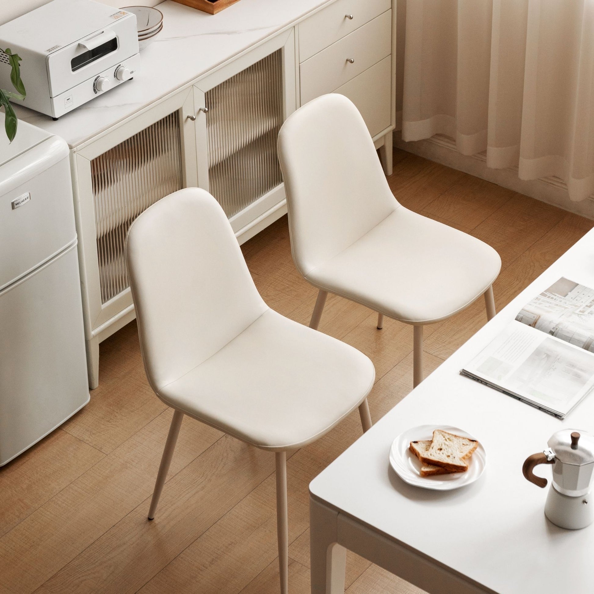 Two white chairs in a room with a table and kitchen appliances.