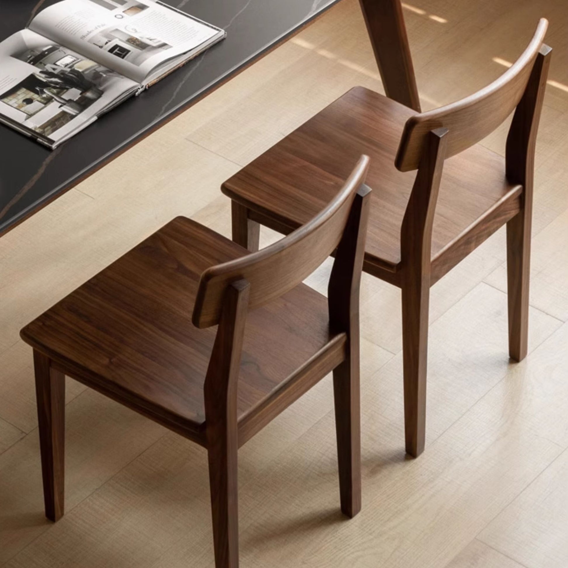 Two wooden chairs in front of a table with an open book on a light wood floor.