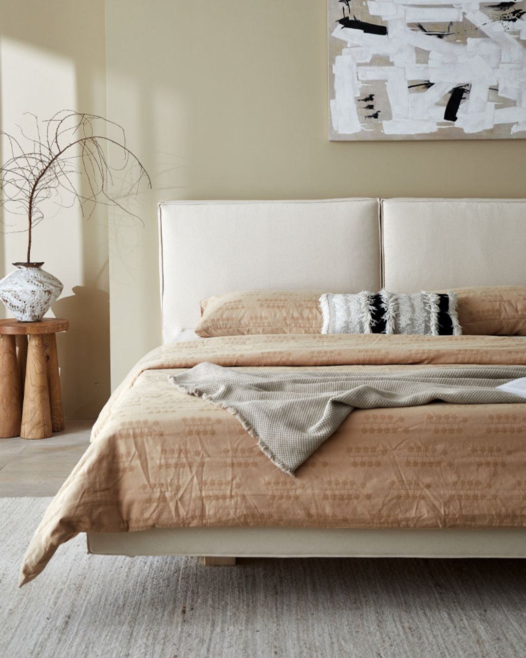 A modern bed with a tall off-white upholstered headboard, dressed in beige textured bedding and a gray throw blanket, set in a minimalist bedroom with a wooden side table and abstract wall art.