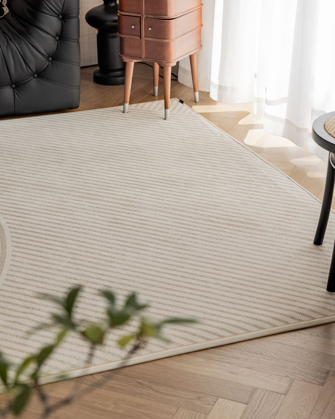 A cream-colored area rug with a subtle striped pattern, styled in a modern living room setting on a light wood herringbone floor, with a tufted sofa, a mid-century side table, and sheer curtains in the background.