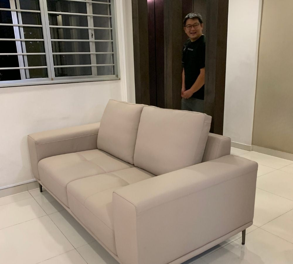 A light beige leather two-seater sofa in a modern living room setting, with a man standing in a doorway behind it.