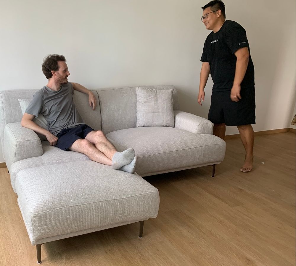 A man in a gray t-shirt and navy shorts sits on a light beige sectional sofa, while another man in a black t-shirt and shorts stands nearby in a room with light wood floors and off-white walls.