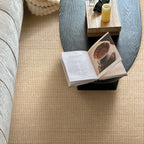 A top-down view of a textured beige woven carpet in a modern living room setting, surrounding a gray sofa and a dark wood coffee table with an open book and a yellow candle.