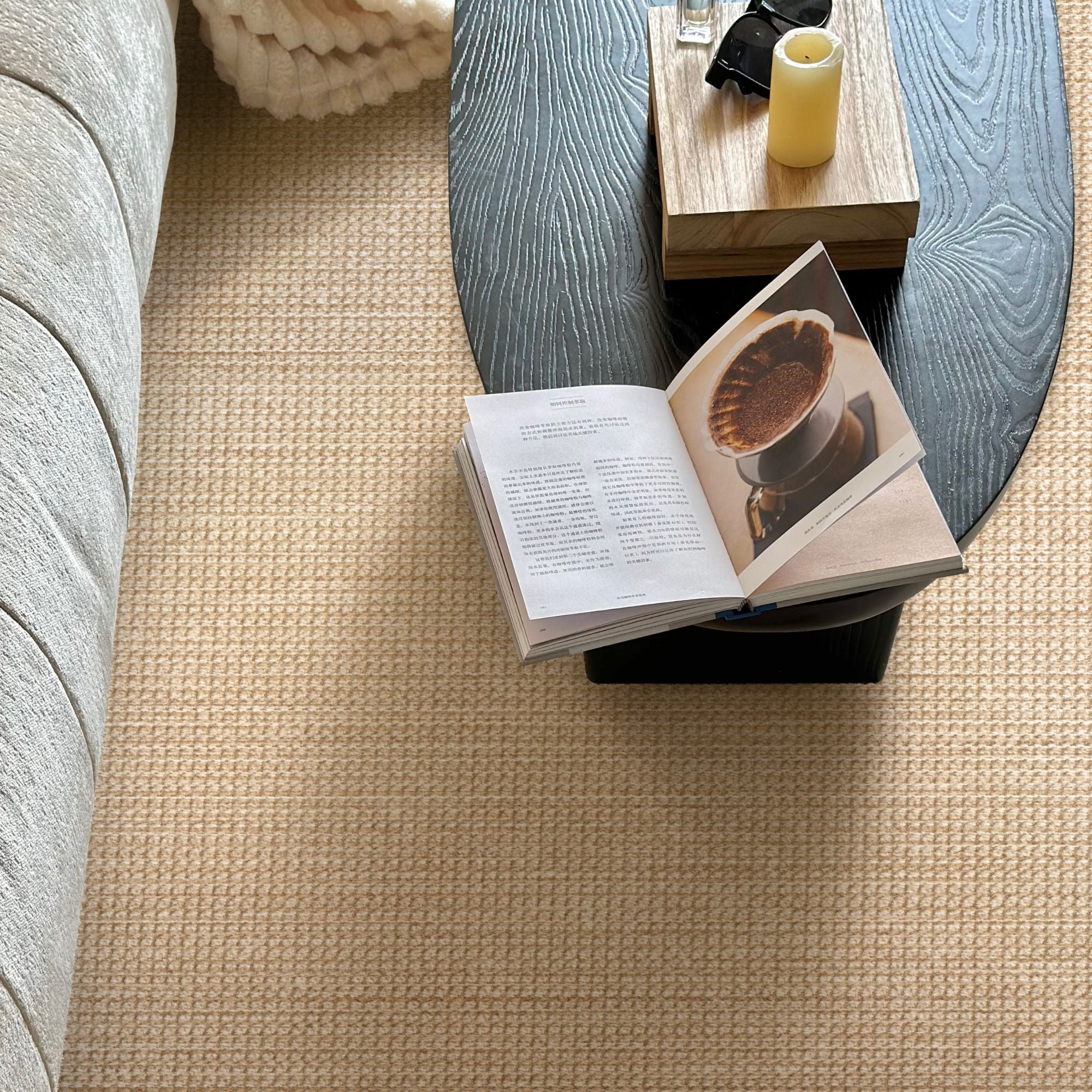 A top-down view of a textured beige woven carpet in a modern living room setting, surrounding a gray sofa and a dark wood coffee table with an open book and a yellow candle.