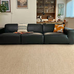 A modern black leather sectional sofa in a living room setting, featuring two decorative throw pillows and a folded blanket, set on a textured beige woven rug.