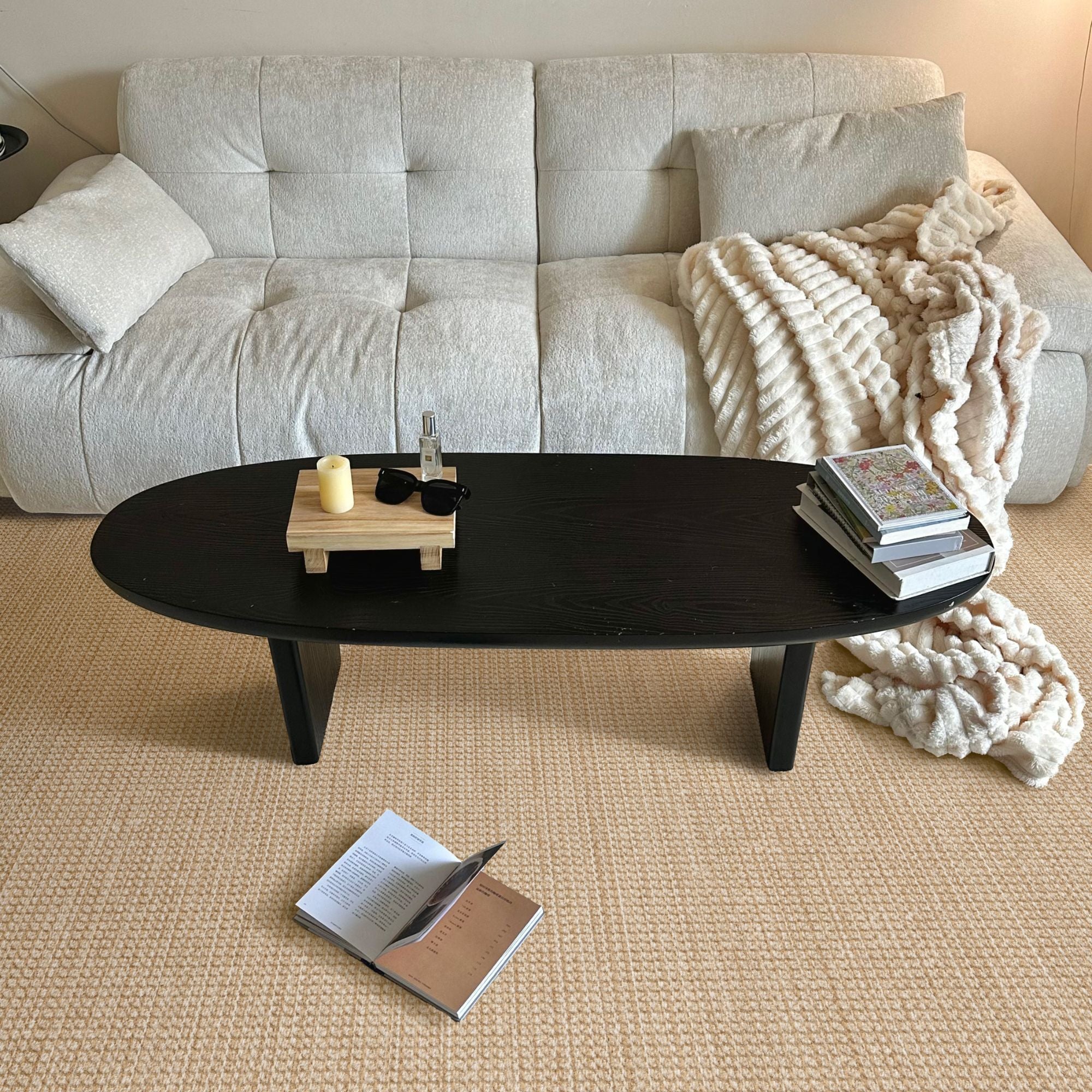 A modern low-profile oval coffee table with a glossy black finish, styled in a cozy living room setting with a beige sofa, textured rug, and a chunky knit throw blanket.