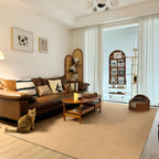 A warm, modern living room featuring a large beige textured area rug as the main flooring. A brown leather sofa with striped and cream pillows sits on the rug next to a small orange tabby cat.