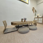 A low wooden table with a live-edge slab top and black metal legs, surrounded by woven floor chairs and round cushion stools in a bright, modern room.