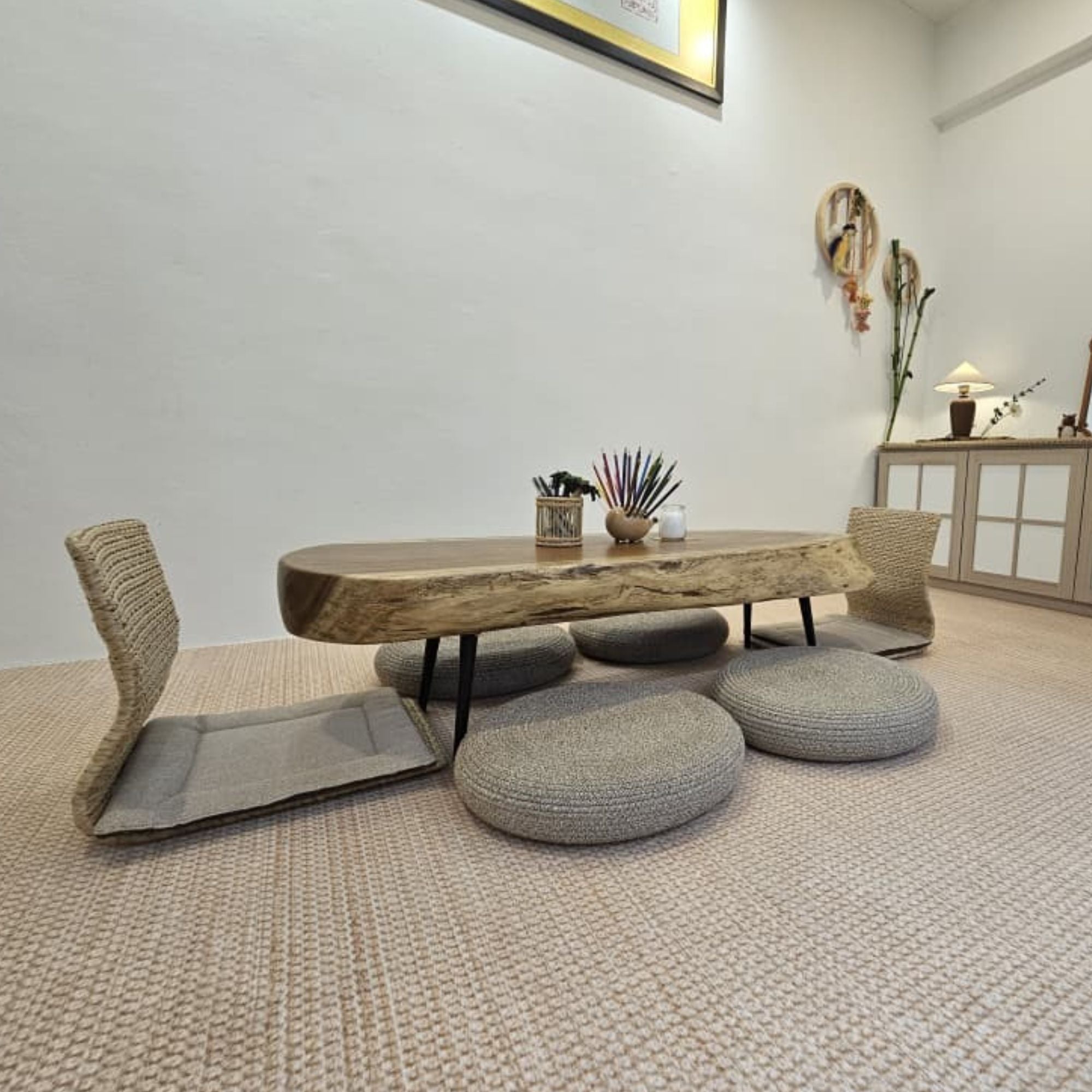 A low wooden table with a live-edge slab top and black metal legs, surrounded by woven floor chairs and round cushion stools in a bright, modern room.