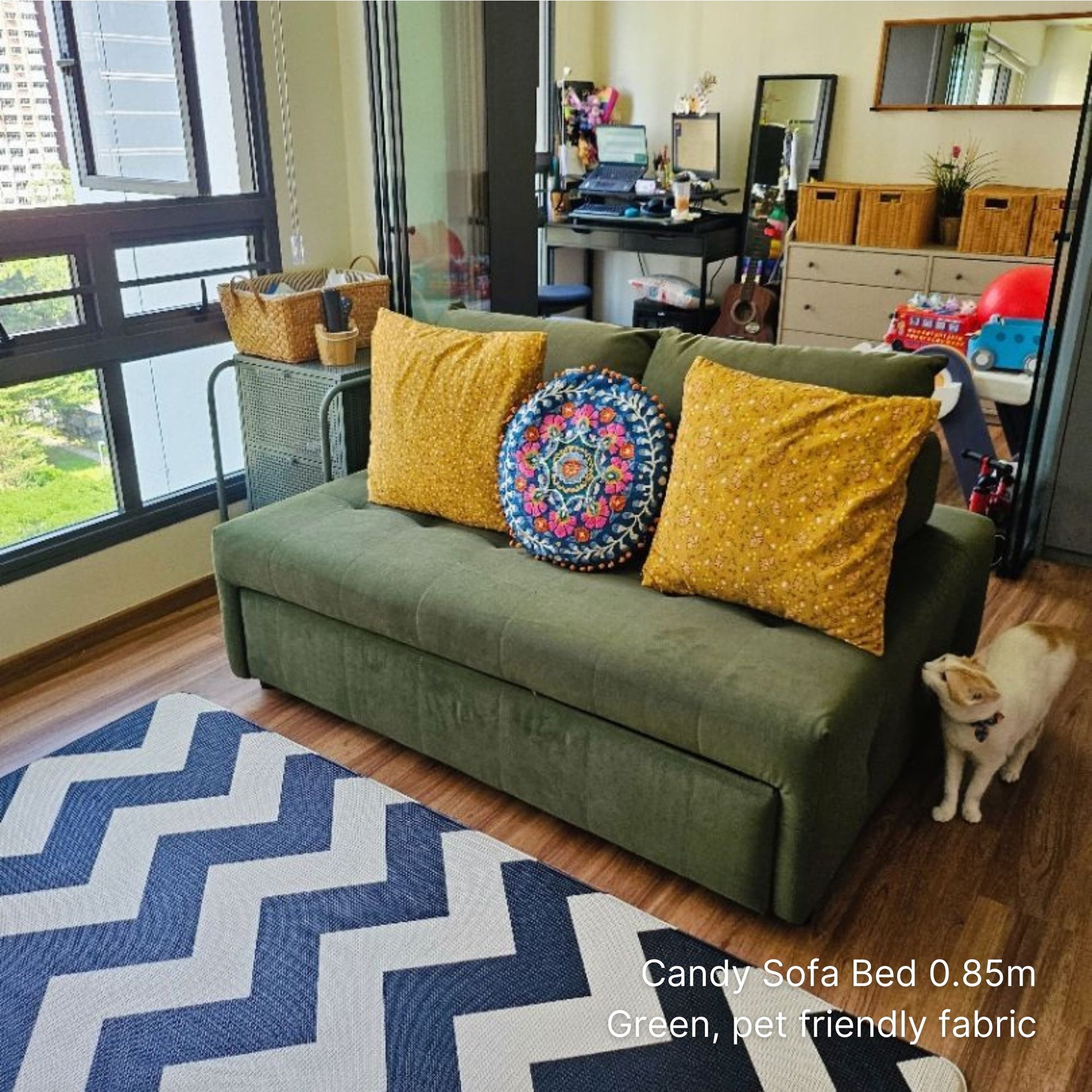 Green sofa bed with yellow cushions and a cat in a room with a desk and windows.