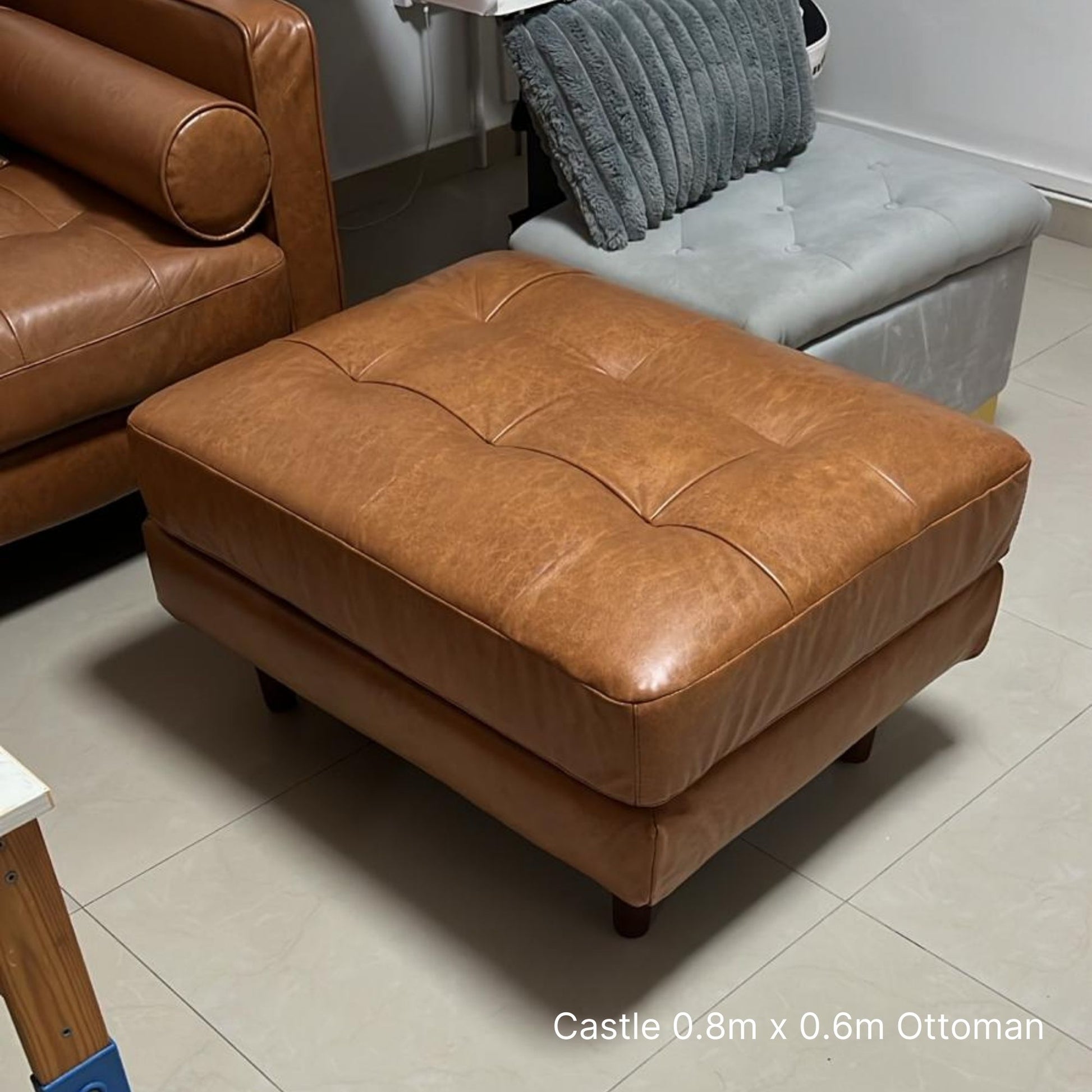 Brown leather ottoman in a room with tiled floor and brown sofa.