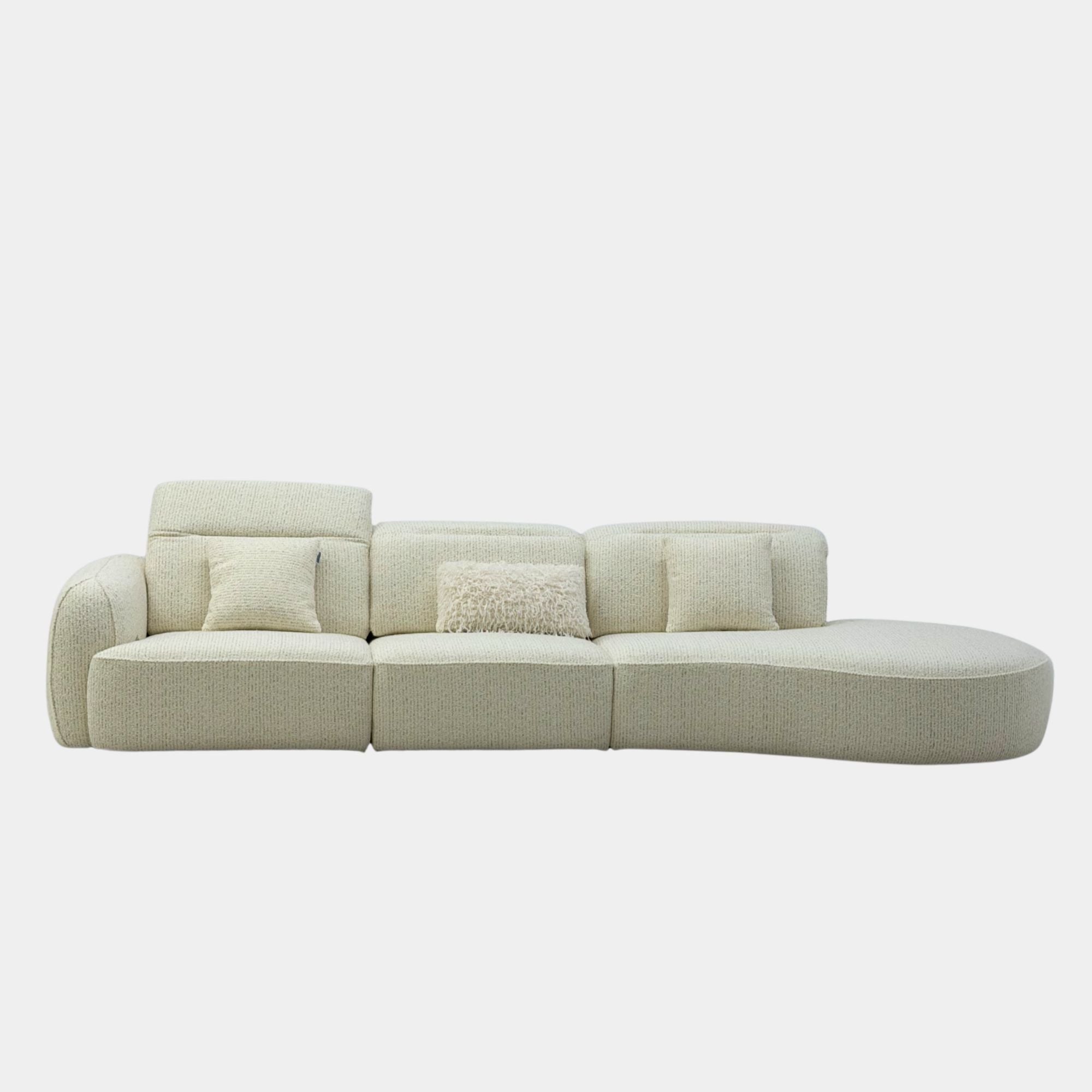A modern curved modular sofa upholstered in a textured cream-beige fabric, featuring two square throw pillows and a central textured lumbar pillow, set against a solid light gray background.
