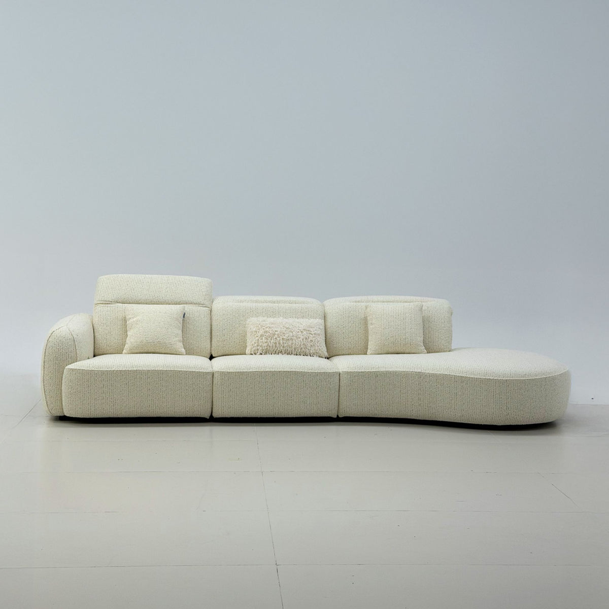 A curved modular upholstered sofa in a warm off-white bouclé fabric, featuring three back cushions, two square throw pillows, and a chaise lounge extension on the right side, set against a minimalist light gray studio background.