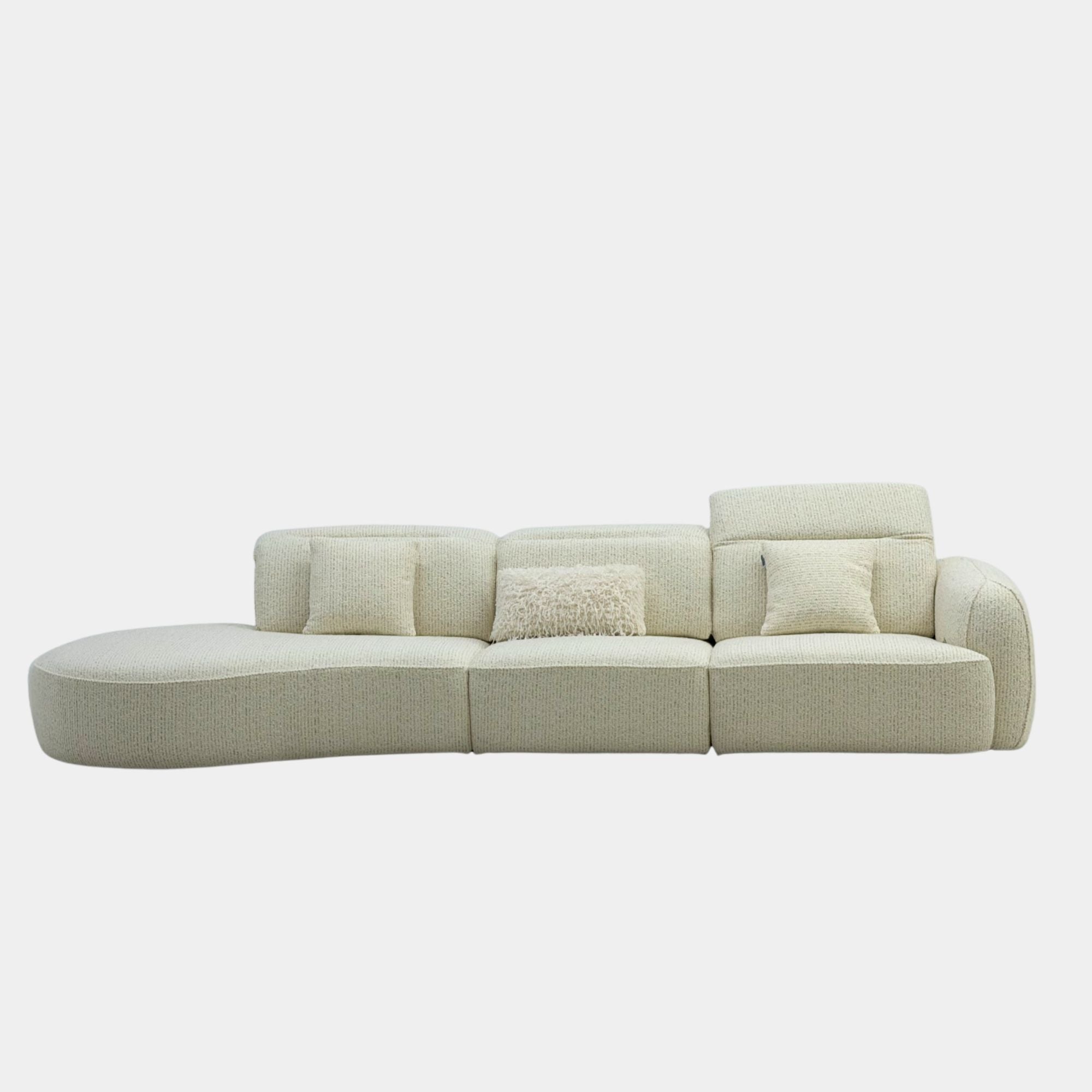 A modern curved modular sofa upholstered in a light beige textured fabric, featuring three matching square throw pillows, including one shaggy accent pillow, set against a plain light gray background.