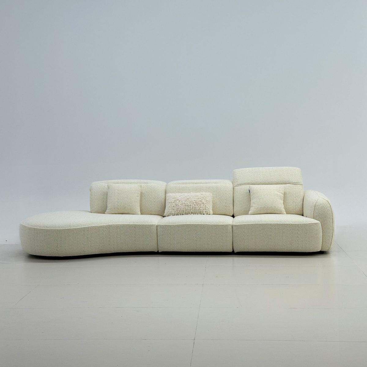 A curved modular upholstered sofa in a light cream bouclé fabric, styled with four matching throw pillows, set against a minimalist light gray studio background.