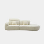 A modern curved modular sofa upholstered in a light beige textured fabric, featuring two matching square throw pillows, shown against a plain light gray background.