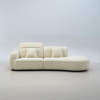 A modern curved modular sofa upholstered in a textured cream fabric, featuring two back cushions, two throw pillows, and a low rounded base, set against a minimalist light gray studio background.