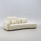 A modern curved upholstered sofa in a light cream or off-white fabric, featuring a low rounded backrest and two matching throw pillows, set against a neutral light gray studio background.