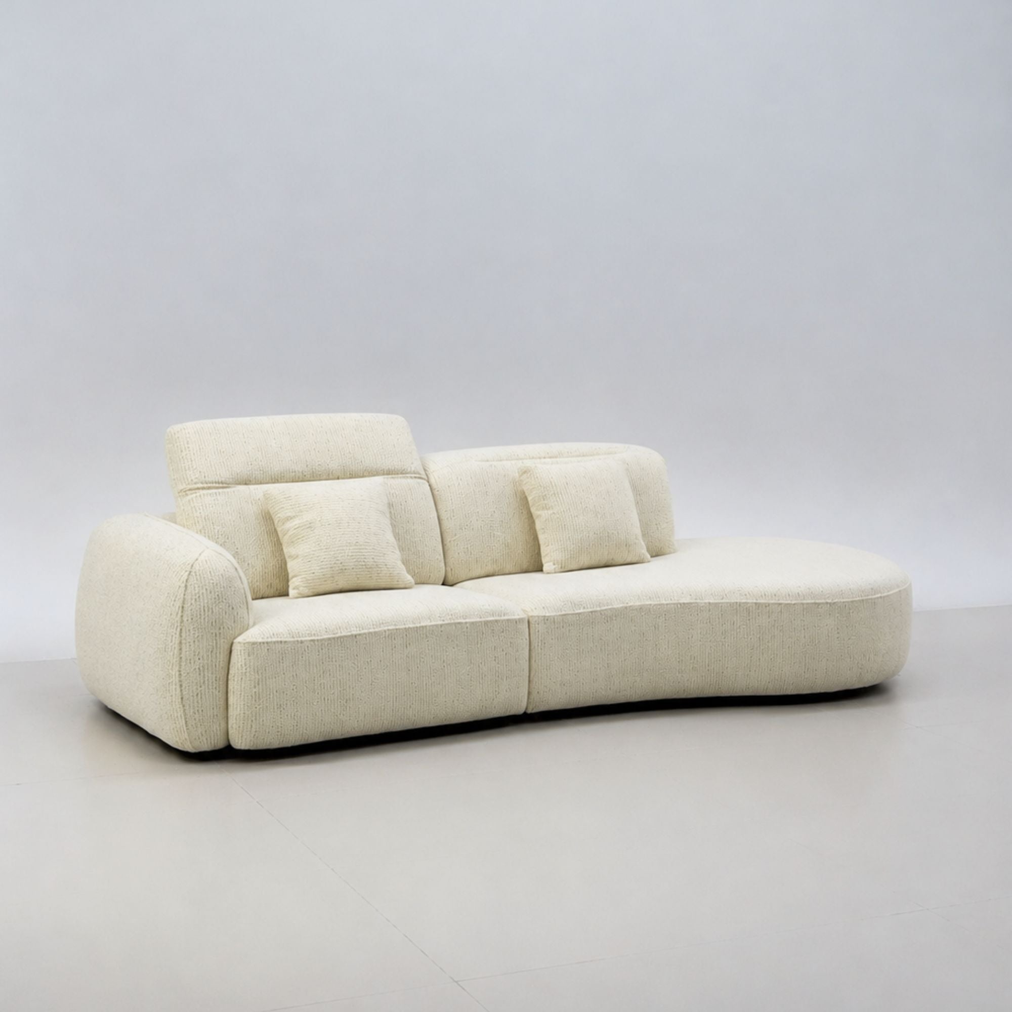 A modern curved upholstered sofa in a light cream or off-white fabric, featuring a low rounded backrest and two matching throw pillows, set against a neutral light gray studio background.