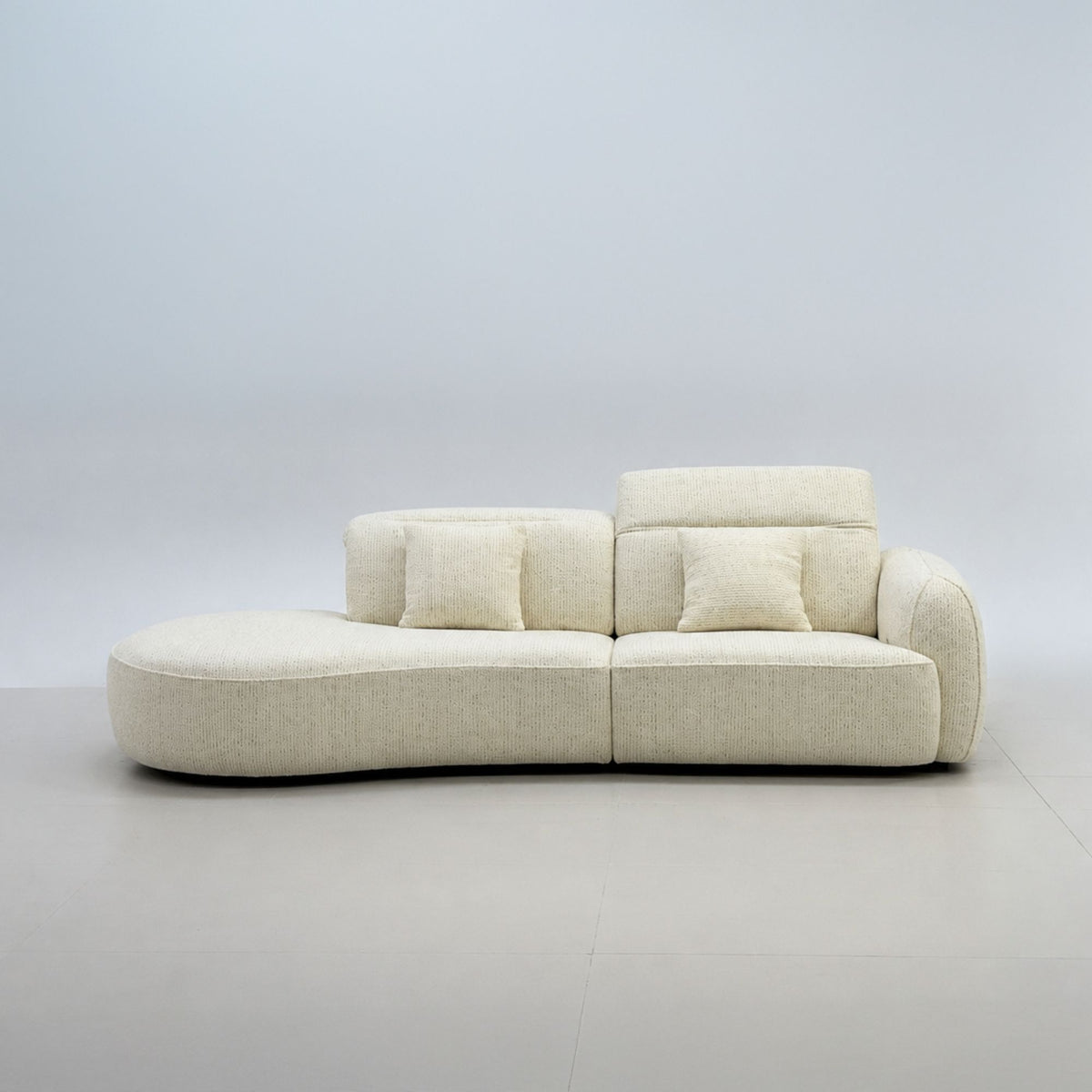 A modern curved modular sofa upholstered in a light beige textured fabric, featuring multiple square back cushions and throw pillows, set against a minimalist light gray studio background.