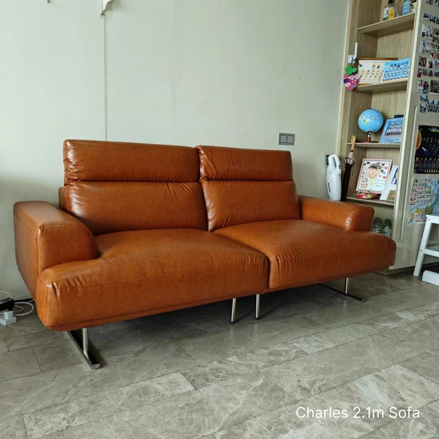 Brown Charles 3 seater 210cm faux leather sofa in a room with a bookshelf and decor items.