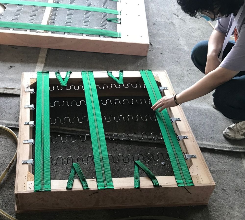 A person is kneeling beside a wooden-framed heating or cooling coil assembly with bright green straps, photographed on a concrete floor. Another similar frame is visible in the background.