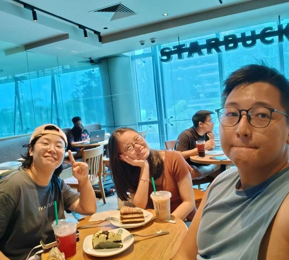 Three friends taking a selfie inside a Starbucks cafe, with food and drinks on the table in the foreground and a large window in the background.