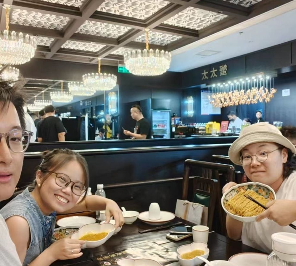 Three people sitting at a restaurant table, with one person on the right holding up a bowl of food. The background shows a restaurant interior with hanging chandeliers, a dark counter, and other diners.