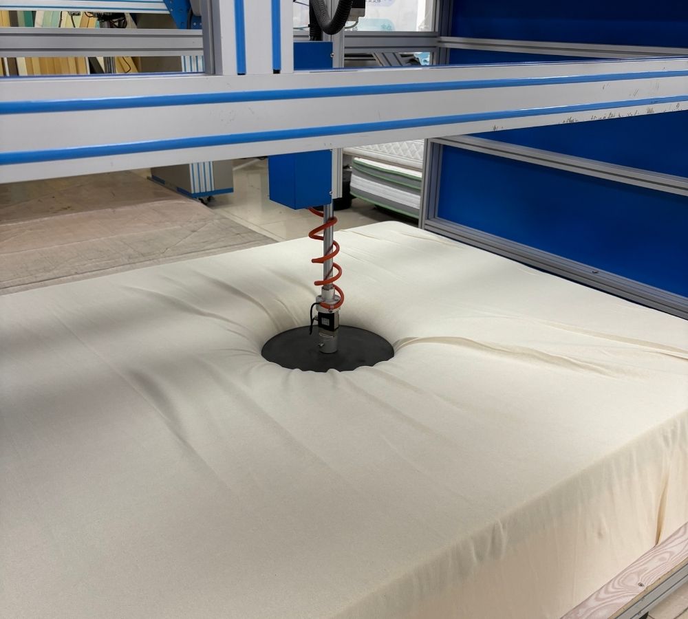 A large cream-colored mattress topper or sheet is being processed by a CNC laser cutting machine, with the laser tip creating a circular cutout in the center. The machine features a blue and white frame, and the background shows an industrial workshop setting.