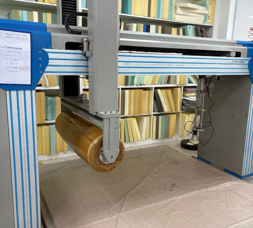 A large industrial sewing or quilting machine with a gray and blue frame, holding a roll of fabric above a flat sheet of matching fabric, set in a storage room with shelves of books in the background.