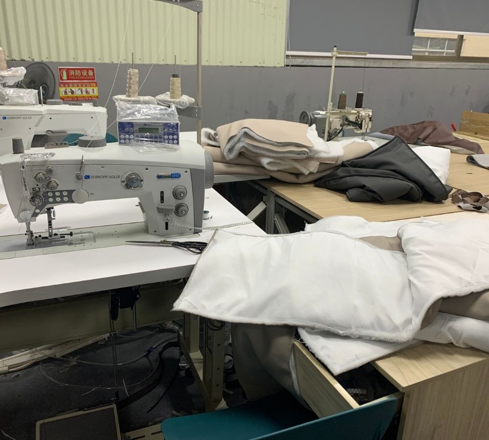 A sewing machine on a worktable in a workshop, with folded white and dark gray fabric pieces stacked nearby and other sewing equipment in the background.