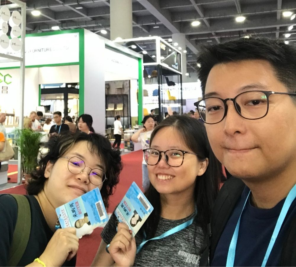 Three people taking a selfie at a trade show or exhibition hall, each holding a blue and white printed card. The background shows booths, other attendees, and exhibition displays.