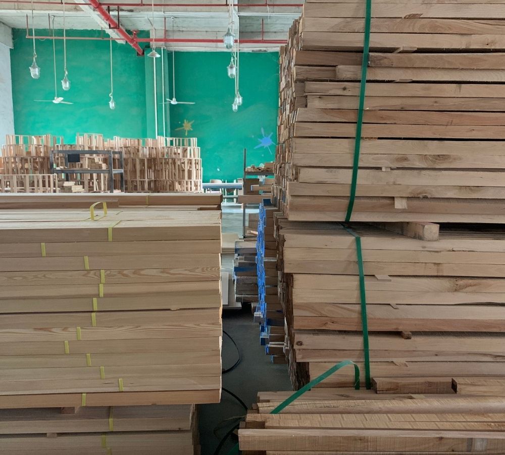 Stacks of light-colored wooden boards and planks secured with straps in a warehouse setting, with a green wall and hanging lights visible in the background.