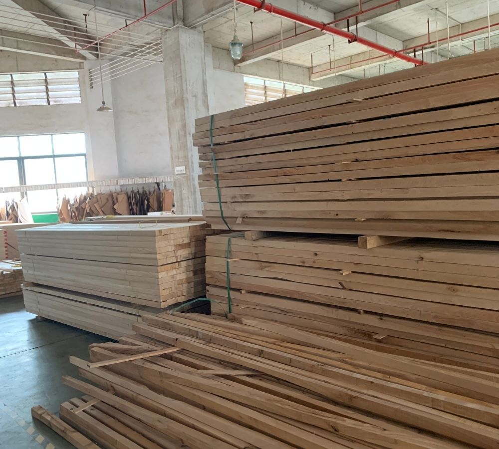 Stacks of natural wood planks and lumber stored in a warehouse or workshop, with concrete pillars, exposed ceiling pipes, and windows in the background.