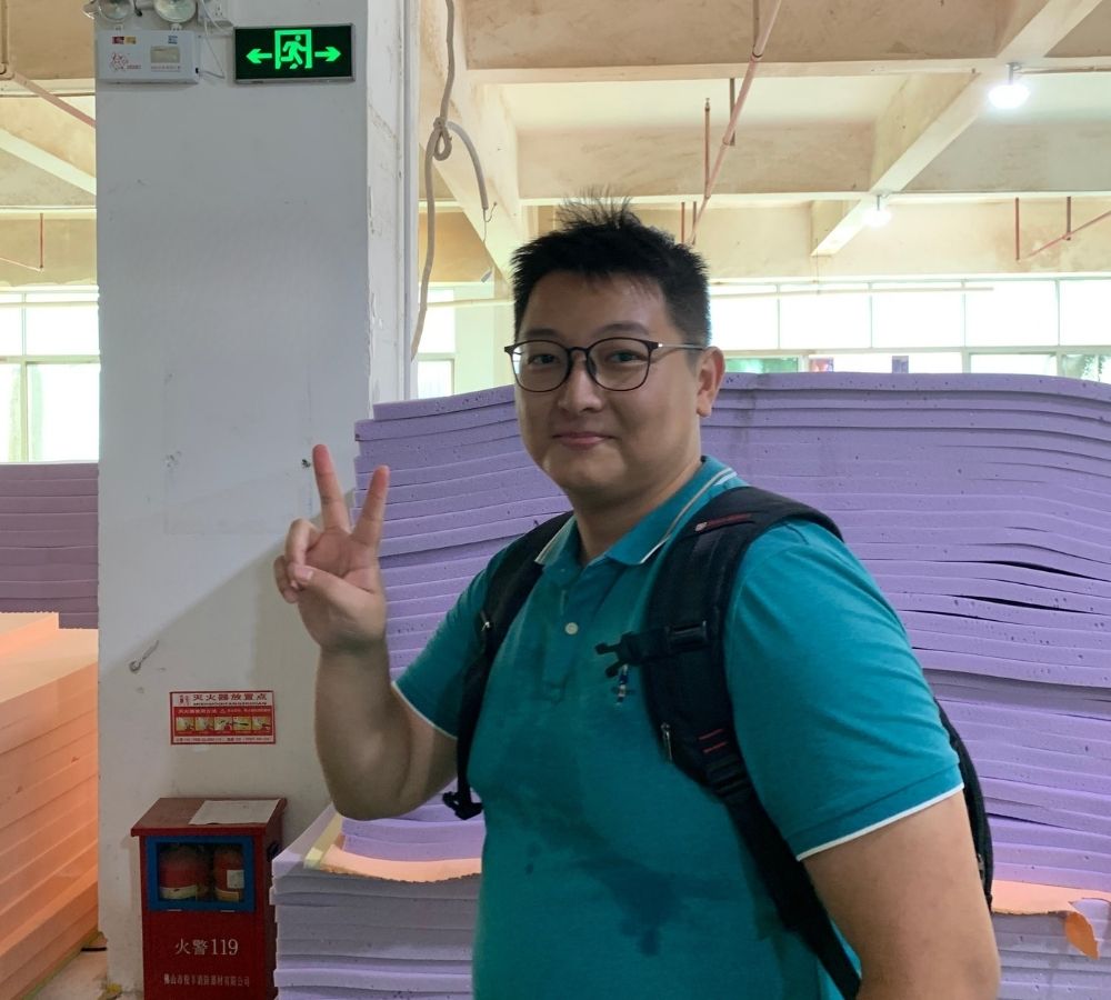 A man wearing a teal polo shirt and a black backpack stands in a warehouse-like setting with stacks of purple foam boards behind him, making a peace sign with his right hand.
