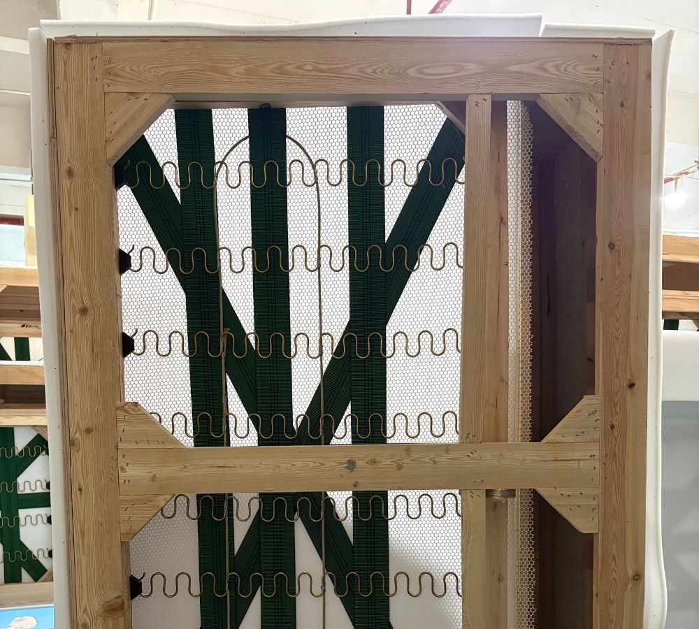 A wooden frame structure with a white mesh backing and dark green wavy support strips, photographed indoors against a light background.