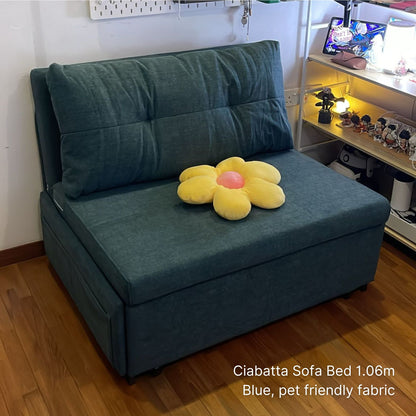 Blue sofa bed with a yellow flower-shaped pillow on a wooden floor, with text indicating dimensions and fabric type.