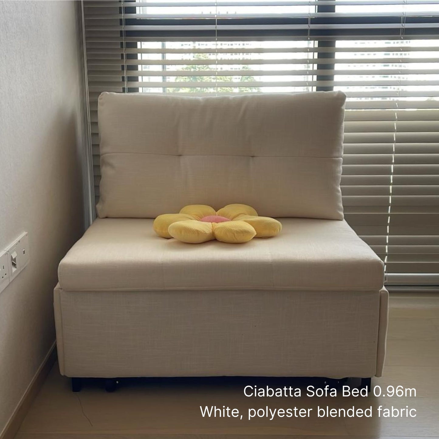 White sofa bed with a yellow flower-shaped pillow in a room with window blinds.