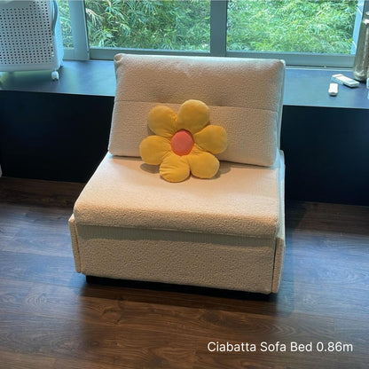 White sofa bed with a yellow flower-shaped pillow in a room with a window and desk.