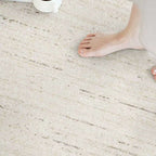 A close-up of a light beige textured rug with subtle gray linear patterns, shown in a lifestyle setting with a person's bare foot stepping onto it.