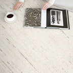 A top-down view of a light beige textured area rug with subtle gray linear patterns, styled with an open book, a white coffee cup, and a person's hands and arms in a white top.