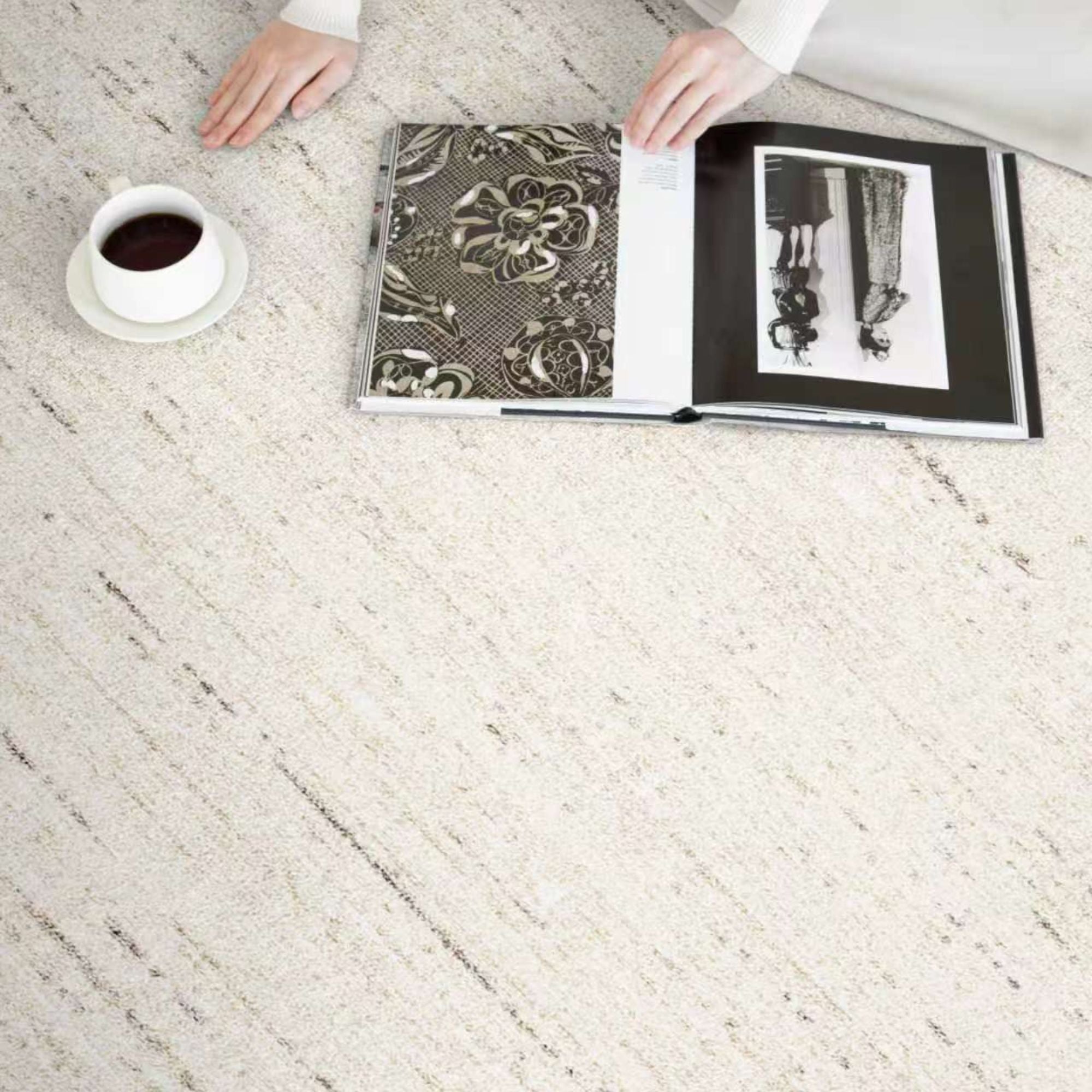 A top-down view of a light beige textured area rug with subtle gray linear patterns, styled with an open book, a white coffee cup, and a person's hands and arms in a white top.