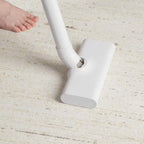 A white cordless vacuum cleaner head attached to a white wand, shown on a light beige textured carpet with a person's bare foot visible at the upper left.