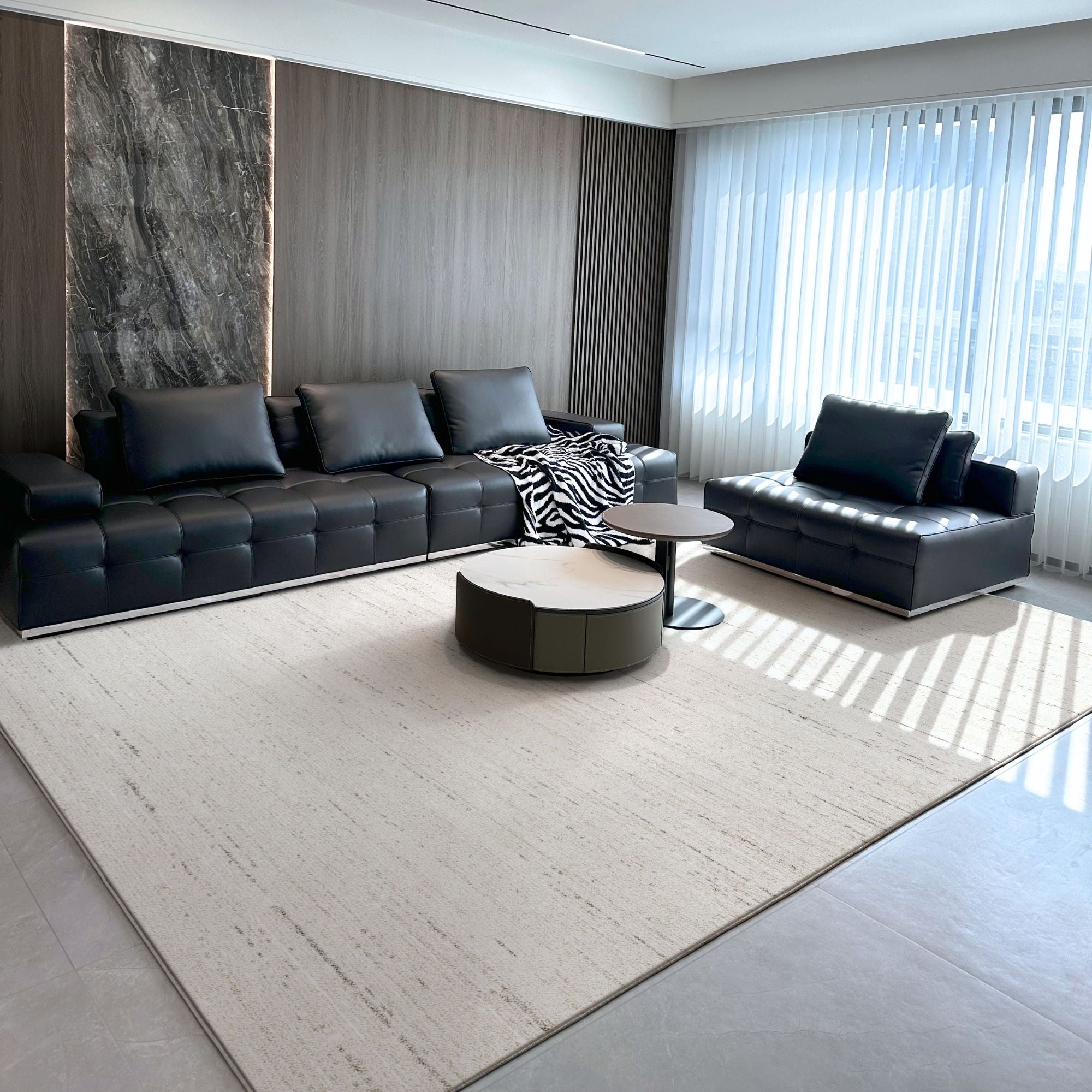 A modern living room featuring a black tufted sectional sofa, a matching black chaise lounge, a round coffee table, and a large light beige textured area rug on a gray tiled floor.