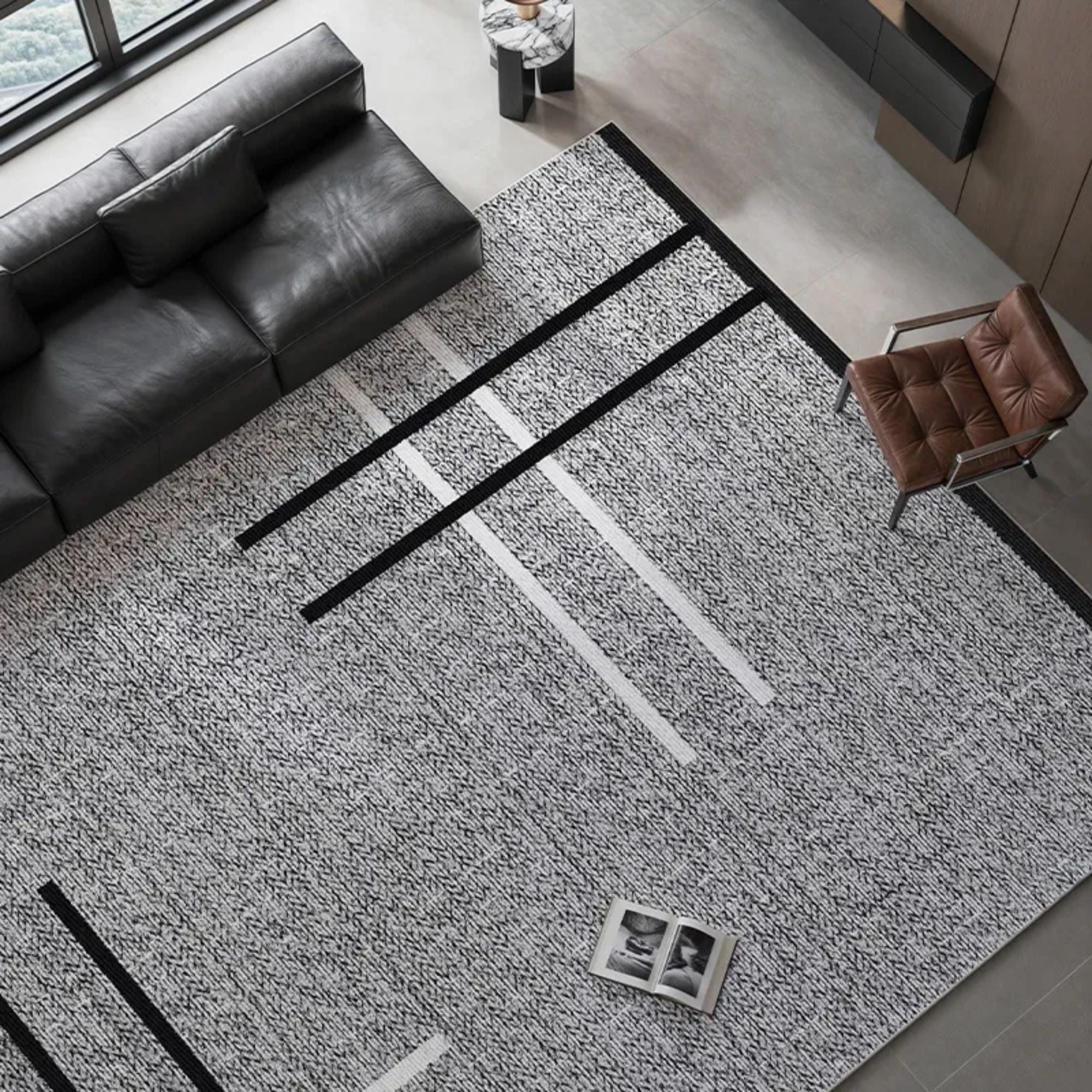 A large modern area rug with a textured black and white pattern and bold black and white linear accents, styled in a contemporary living room next to a dark leather sofa and a brown accent chair.