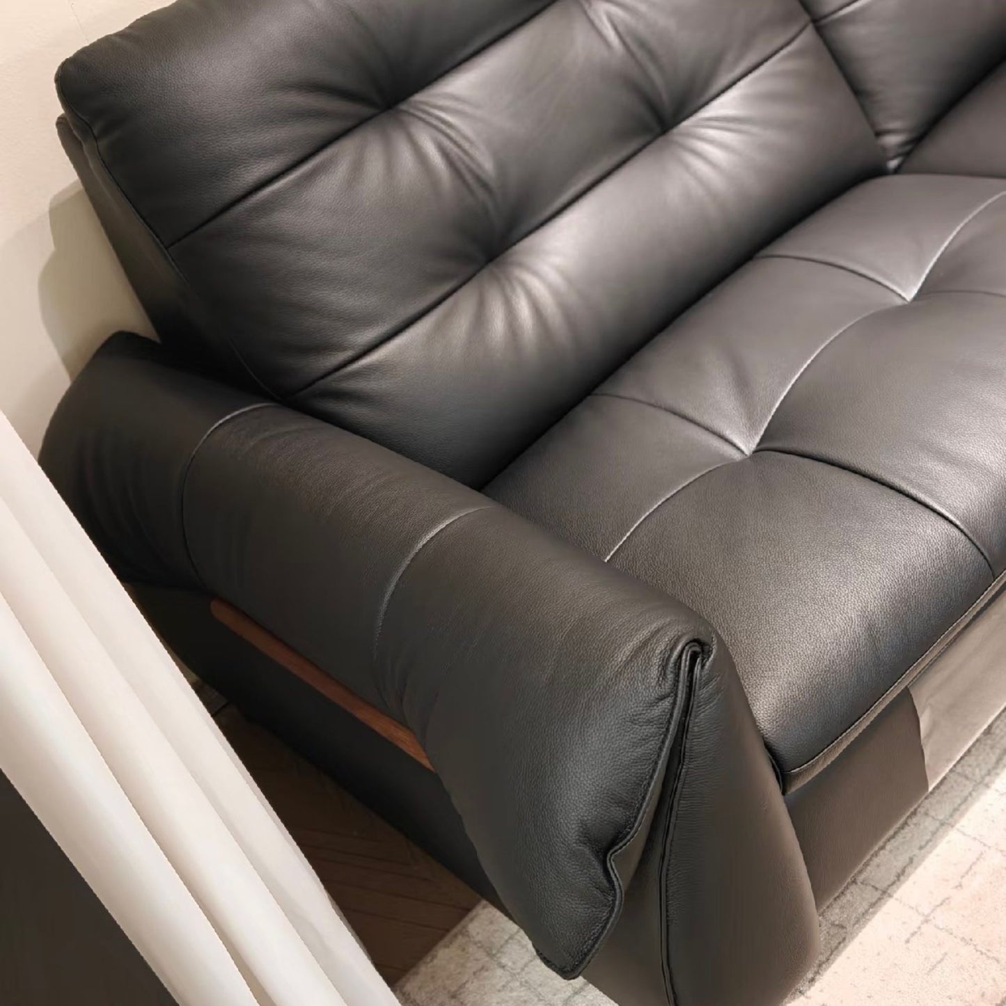 A close-up of a black leather sofa with tufted button details on the backrest and seat, showing the armrest and a small wood accent panel.