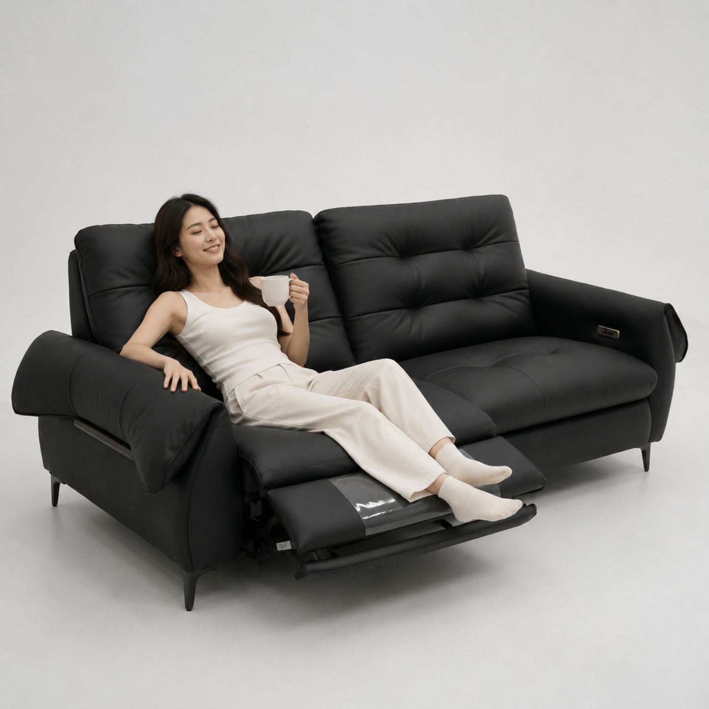 A woman reclines on a black leather sofa with the footrest extended, holding a cup in a bright, minimalist studio setting.