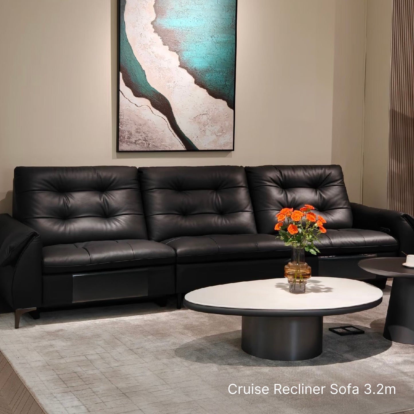 A black leather cruise recliner sofa in a modern living room setting, featuring tufted back cushions, a white oval coffee table with orange flowers, and a large abstract wall art piece above.