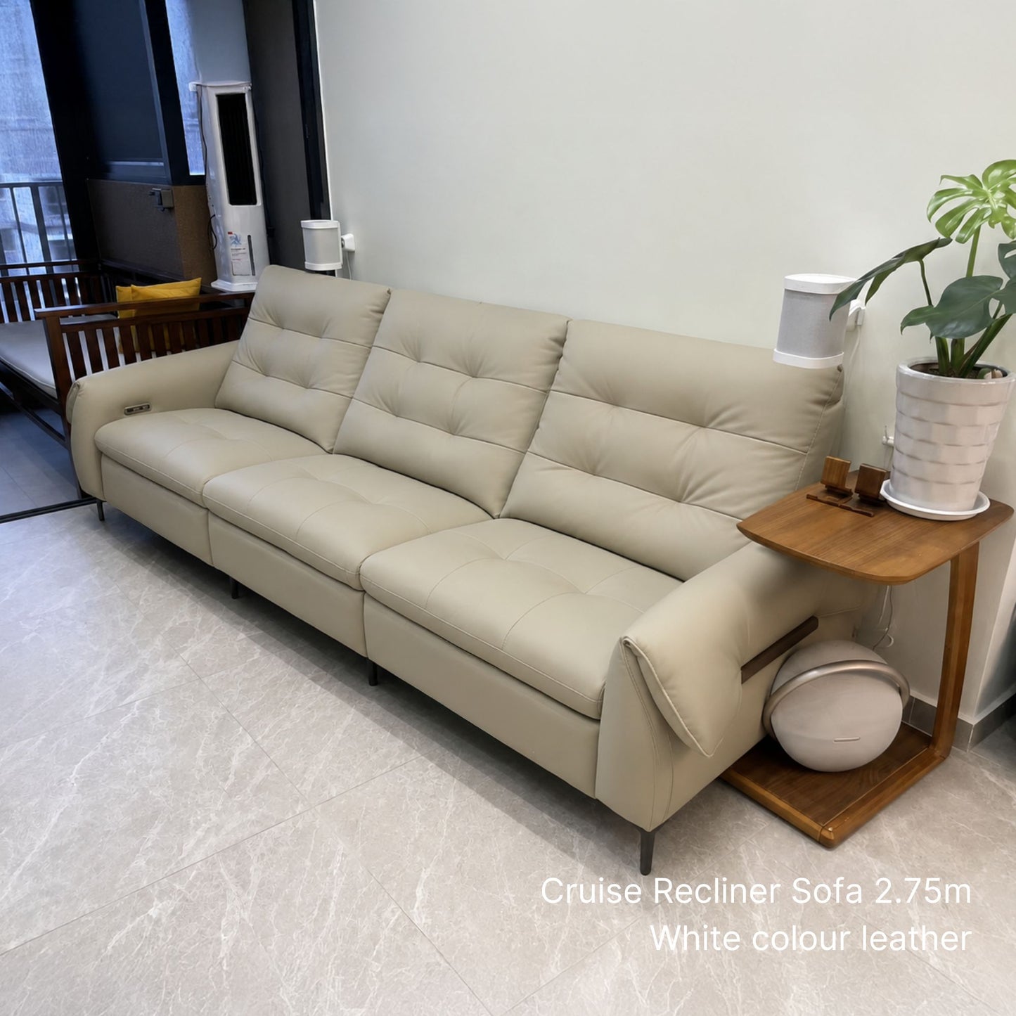 A cream-colored leather cruise recliner sofa in a modern living room setting, paired with a wooden side table holding a lamp and a potted plant.